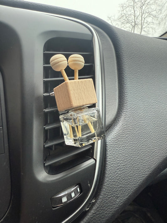 A wooden clip-in car diffuser mounted on a vehicle's air vent, with a glass cube filled with fragrance oil and a dropper bottle.