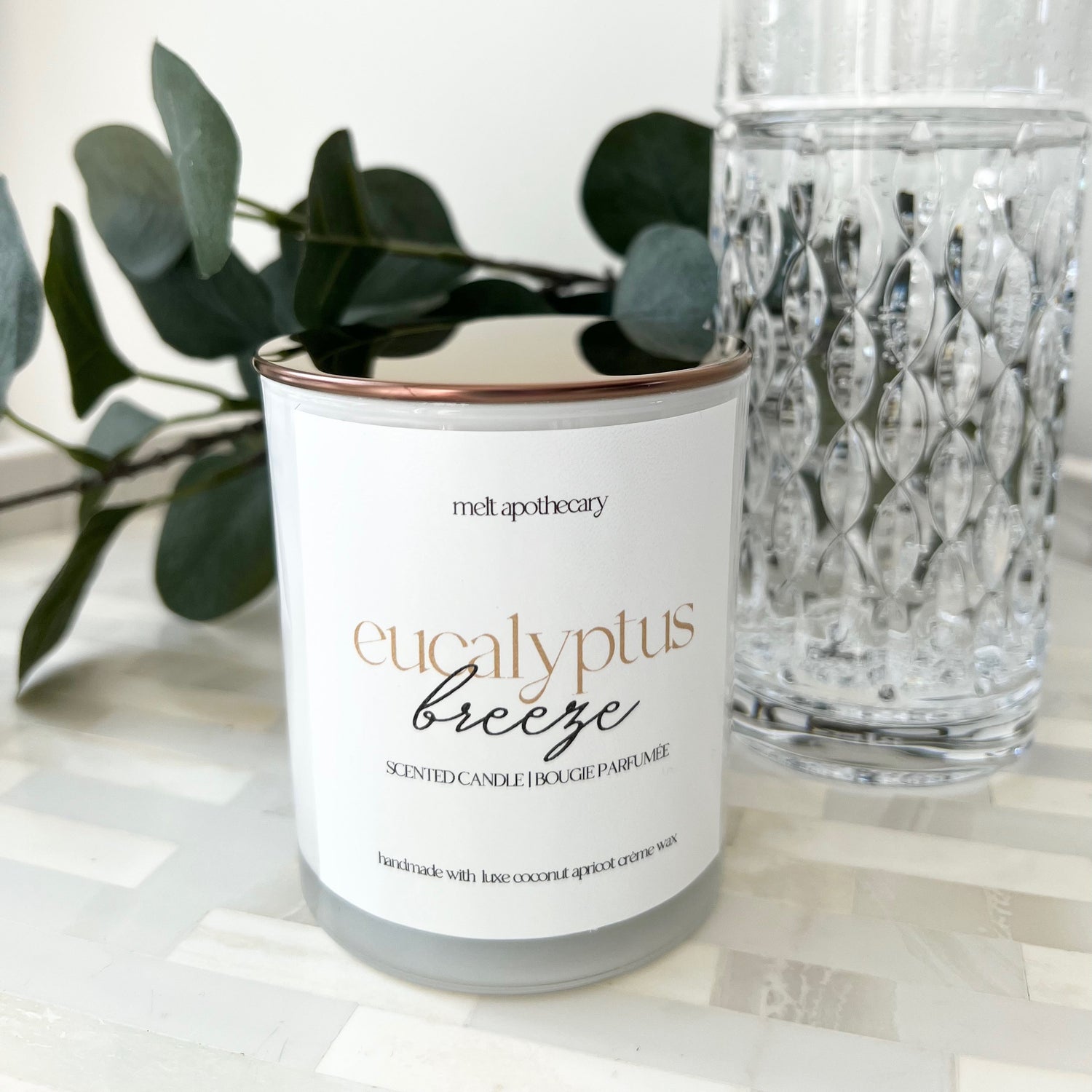 Candle labeled 'eucalyptus freeze' next to a glass and eucalyptus leaves on a white surface