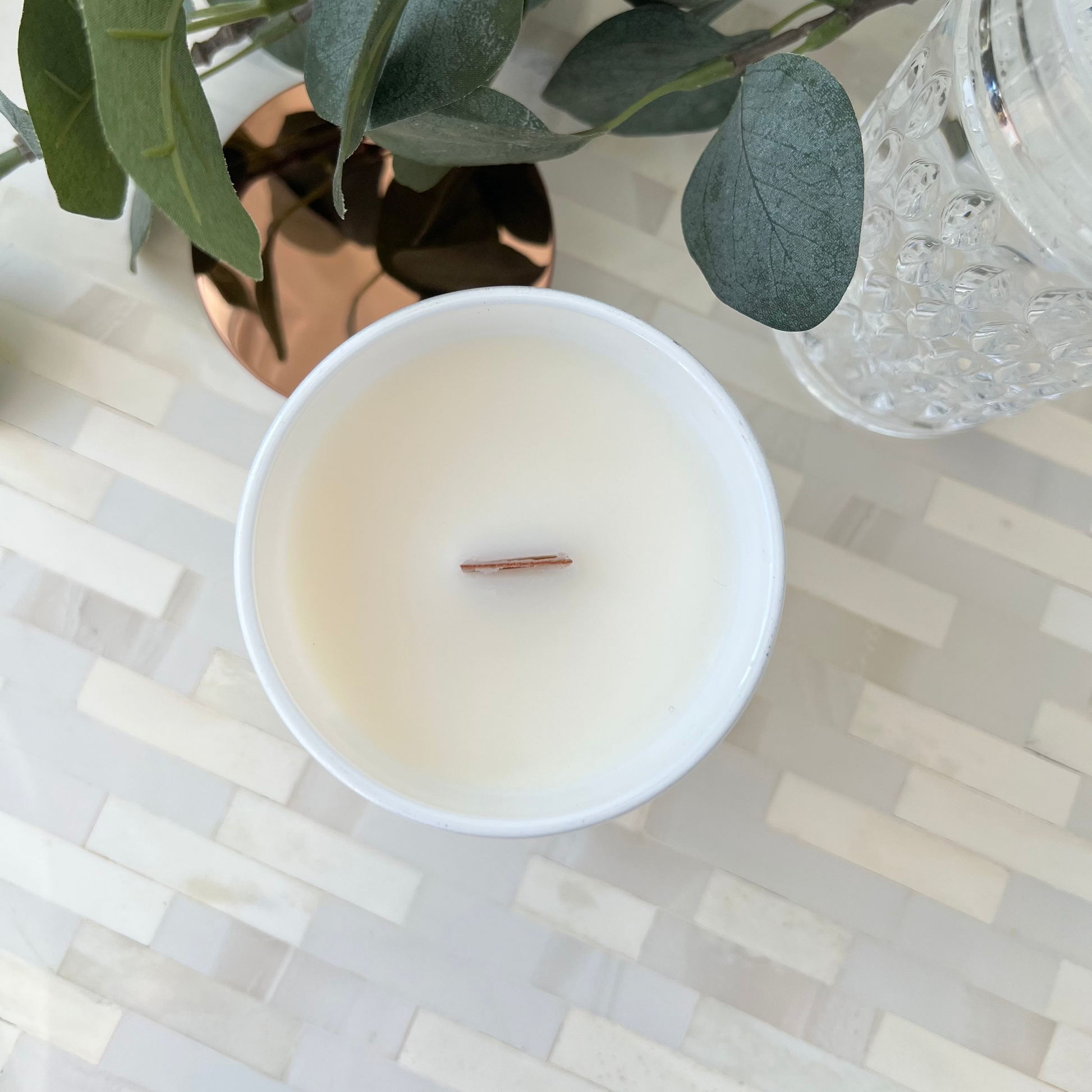 Candle in a white container with a wooden wick, beside green leaves and a vase in the background on a patterned surface