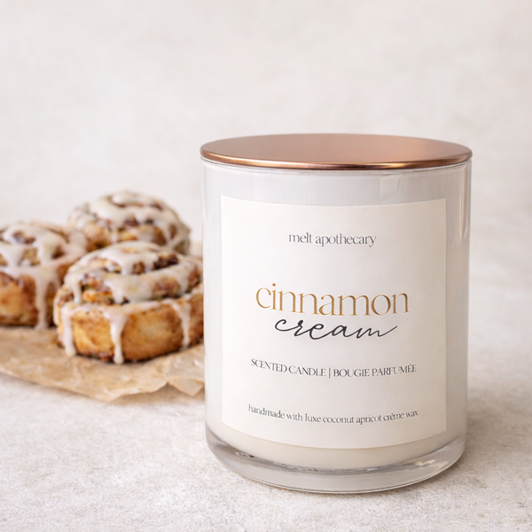Candle labeled 'cinnamon cream' by melt apothecary with cinnamon rolls in the background.