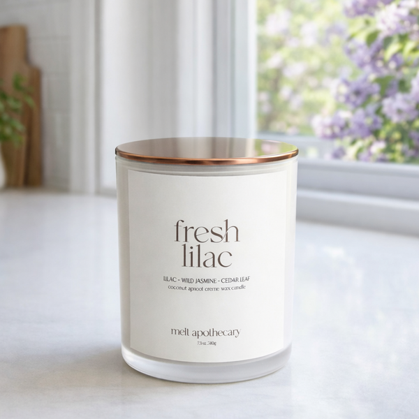Candle labeled 'fresh lilac' on a surface with a window and flowers in the background