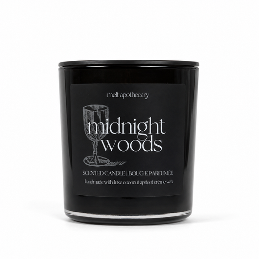 A black glass scented candle jar with a black lid and a black front label reading 'midnight woods' and 'meltpotapothecary'. The label also includes a wine glass illustration and smaller text about coconut and apricot wax.