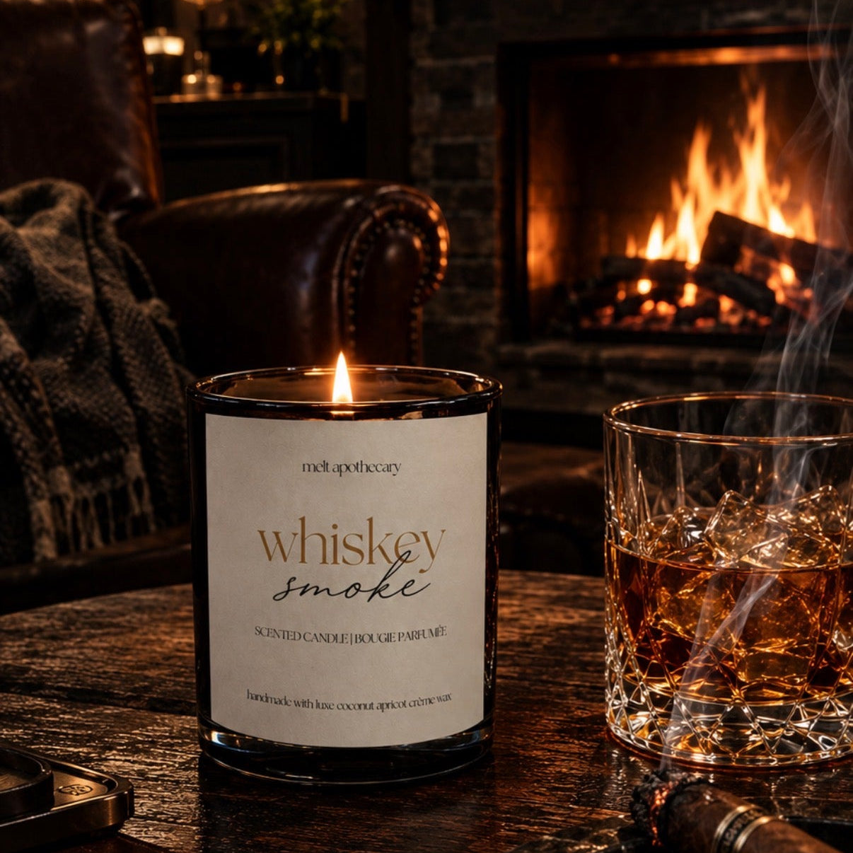 A lit candle labeled 'whiskey smoke' sits on a dark wooden table beside a glass of whiskey and a cigar, with a cozy fireplace and fireplace chair in the background.